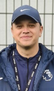 Man wearing a navy blue jacket and cap with a lanyard reading ‘Staff,’ smiling against a background with vertical lines.